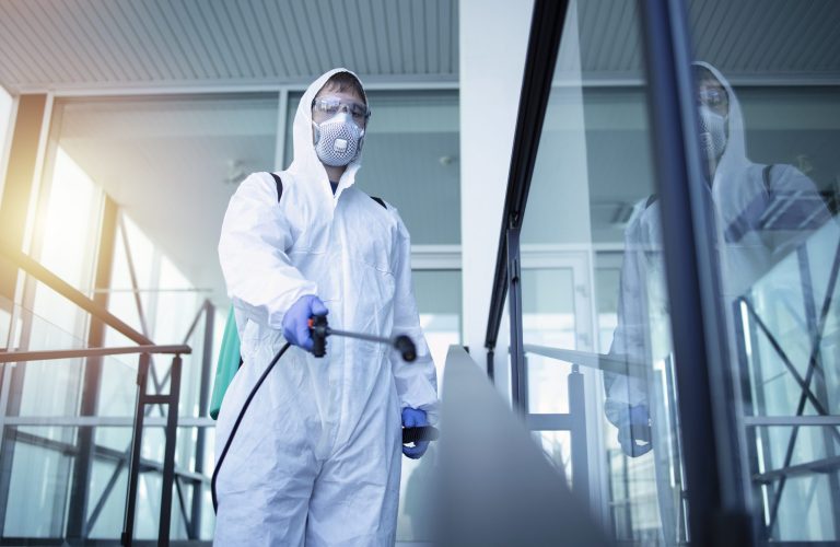 Person in white chemical protection suit doing disinfection of public areas to stop spreading highly contagious corona virus.
